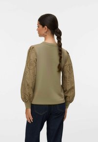 Sudadera caqui con mangas abullonadas que presenta detalles de ojal cortados con láser. Textura suave, cuello acanalado, combinada con jeans de mezclilla oscura.