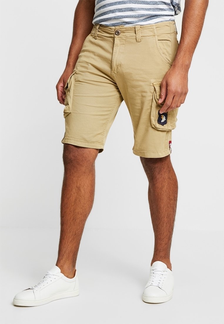 Tan cargoshorts met meerdere zakken, een patch aan de zijkant en een relaxed fit, gecombineerd met witte sneakers en een gestreept shirt.