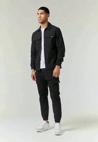 Veste noire avec deux poches avant, portée sur une chemise blanche, associée à un pantalon de jogging noir avec des poches latérales. Des baskets blanches complètent le look.