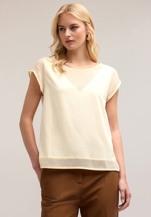 Short-sleeve, light yellow top with a sheer V-panel at the neckline, soft texture, and a relaxed fit. Paired with brown trousers.