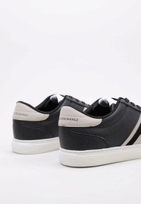 Zapatillas negras con textura de cuero suave, acentos beige en el talón y suela de goma blanca. Presenta dos rayas laterales negras.