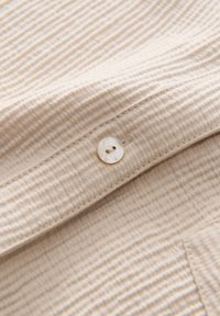 Gros plan d’un tissu côtelé beige avec un petit bouton rond à deux trous cousu sur une patte cousue.