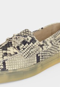 Κοντινή λήψη ενός μοκασίνου slip-on με μπεζ και μαύρο σχέδιο φιδιού και διαφανές λαστιχένιο πέλμα.