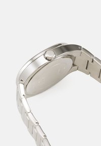 Hopeinen ruostumaton teräksinen kello minimalistisella pyöreällä kotelolla, metallinauhalla ja erottuvalla kruunulla. Liukas, heijastava pinta kiillotetulla viimeistelyllä.