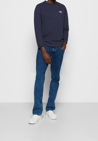 Marinblus med lång ärm och en liten röd och vit grafik på bröstet, i kombination med blå denimjeans och vita sneakers.