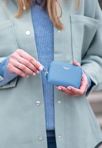 Light blue wallet with a smooth texture, featuring a silver zipper pull and logo. Held in a hand against a soft green outerwear.