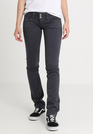Persona que lleva pantalones ajustados gris oscuro con cierre de doble botón y zapatillas negras y blancas, de pie contra un fondo claro y liso.