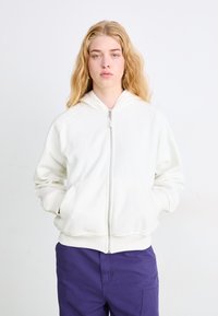 Hanorac alb cu fermoar, din material fleece moale, având glugă, buzunare frontale și manșete cu nervuri. Asortat cu pantaloni mov.