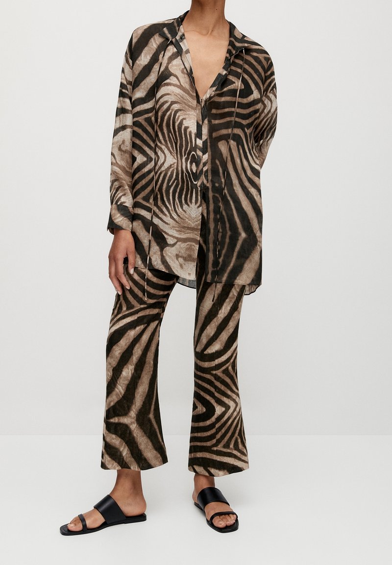 Personne portant un haut ample à manches longues boutonné et un pantalon large avec un motif zèbre marron et noir, ainsi que des sandales slides noires.