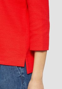 Pullover rosso con texture, maniche a tre quarti, caratterizzato da spacchi laterali e un orlo diritto. Abbinato a denim blu.