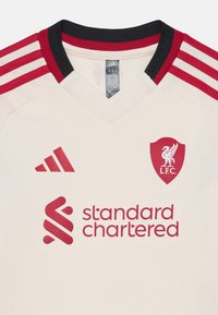 Kremna majica z rdečimi detajli, V-izrez, tri rdeče črte na ramenih, Adidasov logotip, emblem Liverpool FC in "Standard Chartered" v rdeči barvi.