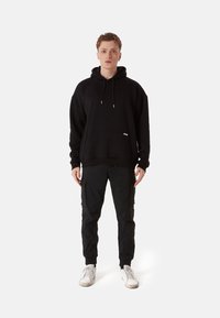 Black hoodie with drawstrings and front pocket, paired with black cargo pants featuring side pockets and ribbed cuffs, worn with white sneakers.