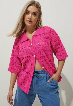 Femme portant un cardigan à manches courtes en maille ajourée rose vif avec des boutons dorés et un pantalon bleu ample à cordon, posant la main dans la poche.