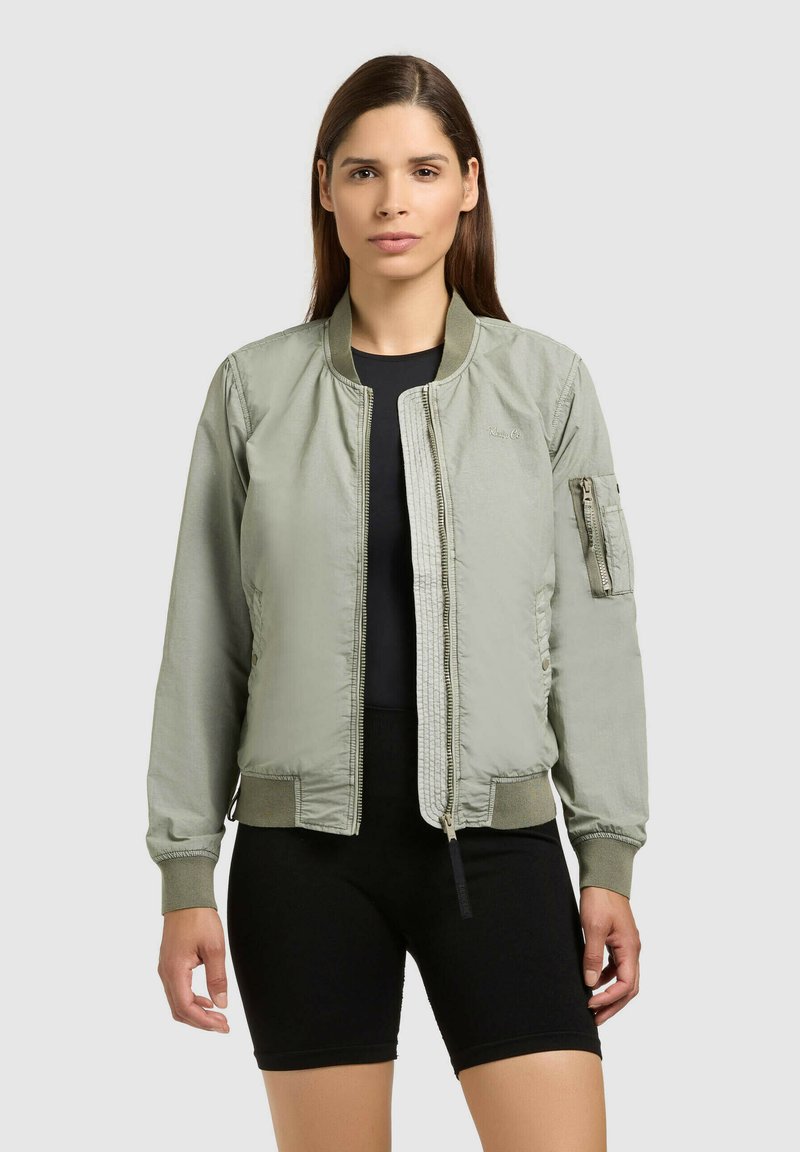 Femme portant un bomber vert clair sur un t-shirt noir et un short de cyclisme noir, debout devant un fond clair uni.