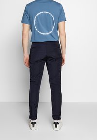 Blå t-shirt med ett cirkulärt vitt mönster på ryggen, tillsammans med mörkblå byxor och vita sneakers med svarta accenter.