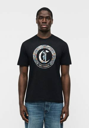 Bărbat purtând tricou negru cu cerc colorat de șerpi și textul „Just Cavalli”, asortat cu blugi albaștri, stând pe un fundal simplu.