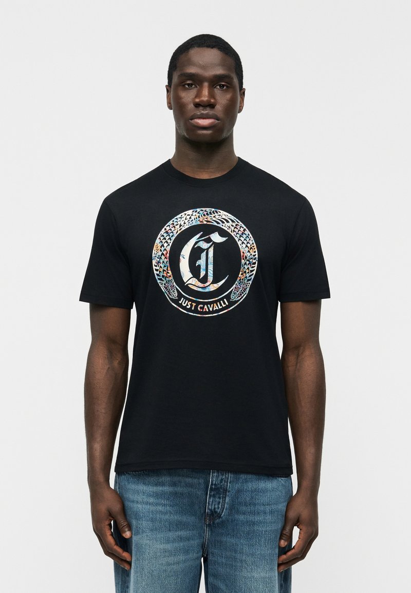 Άνδρας με μαύρο μπλουζάκι που έχει πολύχρωμο κύκλο με φίδι και το κείμενο "Just Cavalli", συνδυασμένο με μπλε τζιν, στέκεται μπροστά σε απλό φόντο.