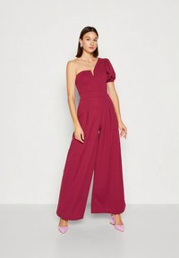 Μπορντό ολόσωμη φόρμα με έναν ώμο, εφαρμοστό μπούστο, βαθύ ντεκολτέ V, φαρδιά γραμμή στο παντελόνι και πτυχώσεις στη μέση, συνδυασμένη με ροζ ψηλοτάκουνα.