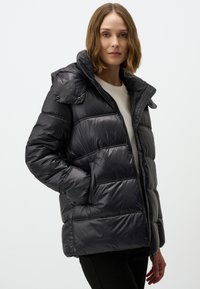 Veste noire matelassée avec une fermeture éclair à l'avant et une capuche amovible. Présente des accents en boutons et des sections matelassées pour plus de texture et de chaleur.