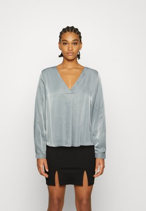 Blusa de seda cinza de mangas compridas com decote profundo em V e costuras franzidas, combinada com uma saia mini preta com duas fendas frontais.