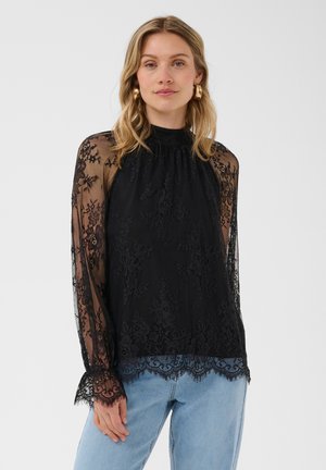 Kvinde iført en langærmet sort blondebluse med gennemsigtige ærmer og blå jeans, stående mod en ensfarvet lys baggrund.