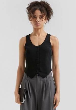 Zwarte mouwloze blouse met een V-hals en zes knopen aan de voorkant, met een aansluitende taille en een uitlopende zoom. Gemaakt in combinatie met grijze broeken en een kleine clutch.