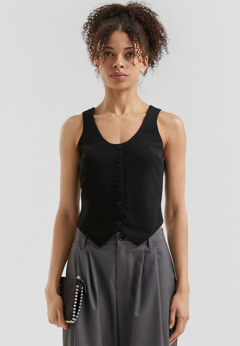 Schwarzes ärmelloses Shirt mit V-Ausschnitt und sechs vorderen Knöpfen, mit einer taillierten Taille und ausgestelltem Saum. Getragen mit grauen Hosen und einer kleinen Clutch.