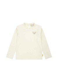 Camicia a maniche lunghe color crema con colletto arricciato e fasce sulle spalle, che presenta una toppa a forma di orso sul petto e un'etichetta sul lato inferiore.