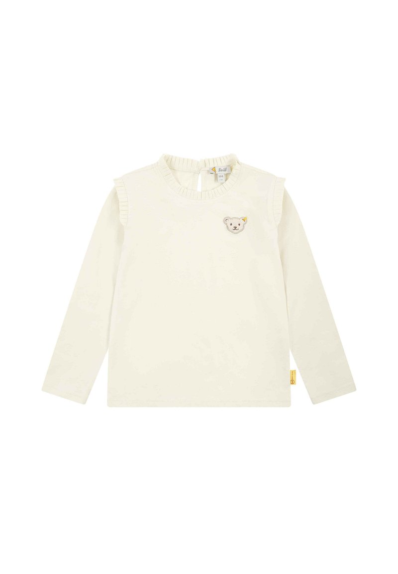Camicia a maniche lunghe color crema con colletto arricciato e fasce sulle spalle, che presenta una toppa a forma di orso sul petto e un'etichetta sul lato inferiore.