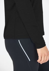 Musta pitkähiha toppi, joka on valmistettu teksturoidusta kankaasta, yhdistettynä tummansinisiin legginsseihin, joissa on valkoinen sivuviiva yksityiskohtana ja kontrastina.