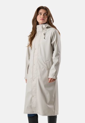 Kobieta ubrana w długi beżowy płaszcz przeciwdeszczowy z kapturem, zapinany na guziki, z kieszeniami, stojąca na jednolitym białym tle.