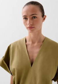 Khaki V-halsjurk met losse, opgerolde mouwen en een zachte textuur. De stof lijkt lichtgewicht met minimale stikseldetails.