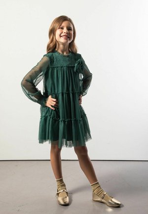 Zielona sukienka z tiulu z długimi przezroczystymi rękawami, warstwowymi falbanami i ozdobą w postaci materiałowego kwiatu. Noszona ze złotymi butami i pasującymi skarpetkami.