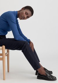 Niebieska żebrowana bluzka z długim rękawem, ciemnoniebieskie dopasowane spodnie oraz czarne skórzane mokasyny z detalem w postaci frędzla. Model siedzi na drewnianym krześle.