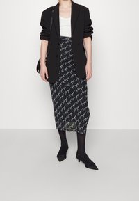 Blazer negro ajustado sobre una camiseta blanca sin mangas, combinado con una falda midi negra con un patrón manuscrito en blanco. Medias negras y tacones de punta.