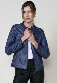 Giacca da moto in pelle blu con design aderente, dettagli trapuntati sulle maniche e due tasche con zip sul petto. Indossata sopra un top bianco.