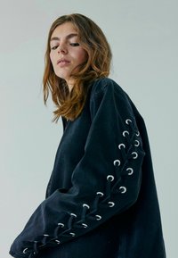 Musta ylijä�ämän paita, jossa on nauhoitusyksityiskohtia hihoissa, metalliset aukot ja punottuja aksentteja. Pehmeä kangas.