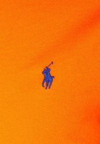Oranžová bavlněná látka s hrubou strukturou, na levé straně s modrou vyšitou námořnickou logem polo hráče.