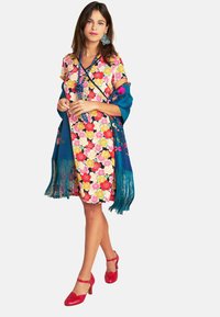 Blumenmusterkleid in Rosa, Gelb und Rot mit kurzen Ärmeln. Blauer Schal mit Fransen, rote Absatzschuhe und blaue Accessoires.