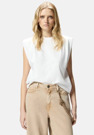 Donna bionda con onde morbide indossa un top bianco senza maniche con imbottiture sulle spalle e jeans beige a vita alta con accessorio intrecciato a nappine.