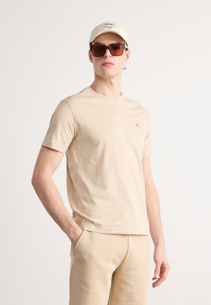 Beige bomulds T-shirt med korte ærmer, parret med matchende beige bukser. Accessories inkluderer en lys beige kasket og mørke solbriller.