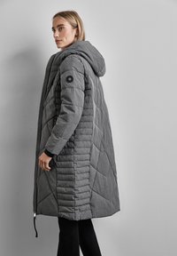 Manteau rembourré gris avec capuche, présentant un matelassage en losanges, une fermeture à glissière et un accent de logo circulaire sur la manche.