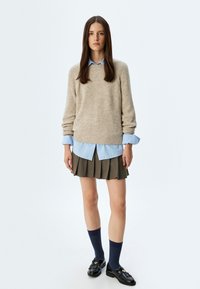 Maglione di lana beige sopra una camicia blu con colletto, abbinato a una gonna a pieghe verde oliva, calze scure e mocassini neri con un accento dorato.