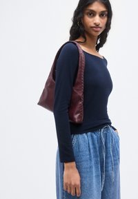 Ένα τσαντάκι ώμου από βουργουνδί δέρμα με πλεκτές λεπτομέρειες, δομημένο σχήμα και μία μονή λουρίδα ώμου, συνδυασμένο με μπλε σκούρο μπλούζα μακριάς μανίκας και τζιν παντελόνι.