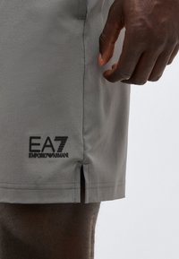 Κοντινό πλάνο γκρι σορτς EA7 Emporio Armani με πλαϊνή σχισμή, που φοριέται από άτομο με σκουρόχρωμο δέρμα, δείχνοντας μέρος του χεριού και του μηρού του.