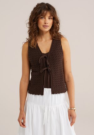 Vrouw met golvend bruin haar draagt een mouwloze donkerbruine gebreide spencer, vooraan gestrikt, over een bruin shirt en een witte rok, staat neutraal.