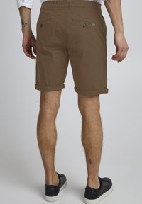Shorts en coton marron avec un ourlet roulé, dotés de deux poches arrière et d'une étiquette de marque discrète sur le côté droit. Portés avec des baskets noires.