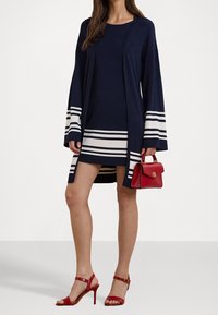 Námorně modrý cardigan s bílými horizontálními pruhy na manžetách, spárovaný s námořně modrými šaty s bílými pruhy. Červená kabelka a odpovídající lodičky.