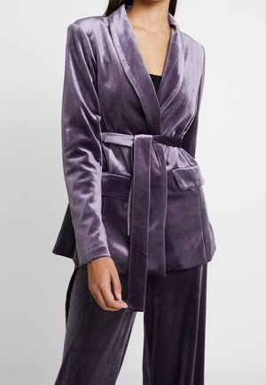 Kobieta ubrana w fioletową aksamitną marynarkę z paskiem i klapowymi kieszeniami oraz pasujące aksamitne spodnie na jednolitym tle.