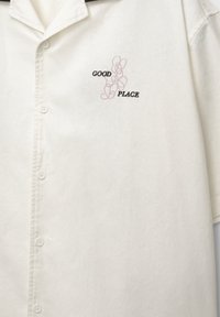 Chemise écrue à manches courtes avec col et un motif indiquant "GOOD PLACE" avec des boucles roses abstraites sur la poitrine.